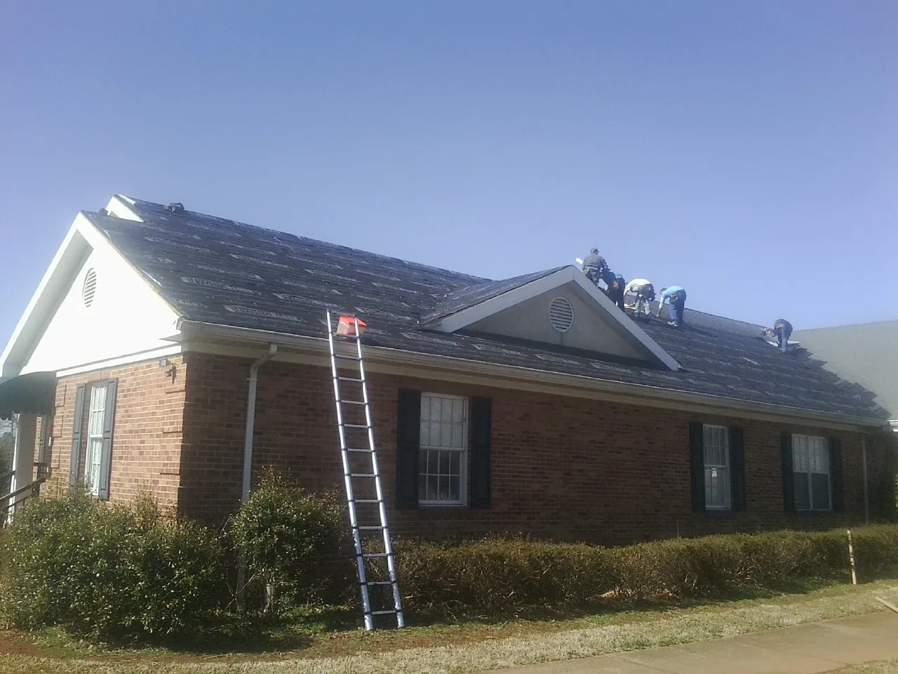 Crew tearing shingles off a brick ranch roof with a ladder set — Emergency Roof Repair work by our crew in Manson